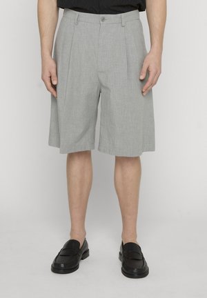 Mann trägt hellgraue Faltenshorts, schwarze Loafers und ein in die Hose gestecktes schwarzes Hemd und steht vor einem schlichten weißen Hintergrund.