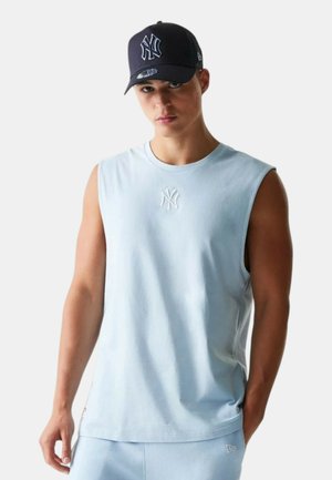 Giovane uomo che indossa un top senza maniche azzurro chiaro e pantaloni abbinati con cappellino blu navy dei New York Yankees, in piedi davanti a uno sfondo semplice.