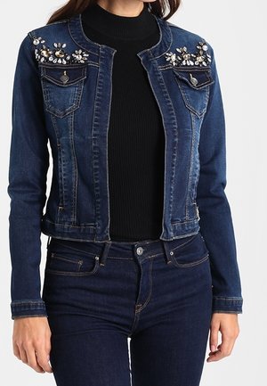 Mujer que lleva una chaqueta vaquera azul oscuro con adornos de gemas en los hombros, combinada con un top negro y jeans azul oscuro.