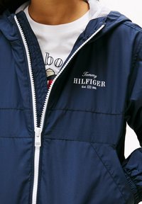 Námořnická lehká bunda s kapucí, bílým zipem a výšivkou loga "Tommy Hilfiger" na hrudi. Má elastické manžety a švy.