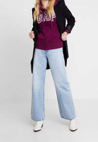 Femme portant un manteau noir, un hoodie violet de GAP, un jean large bleu clair et des bottines blanches, se tenant debout avec les mains ajustant son manteau.