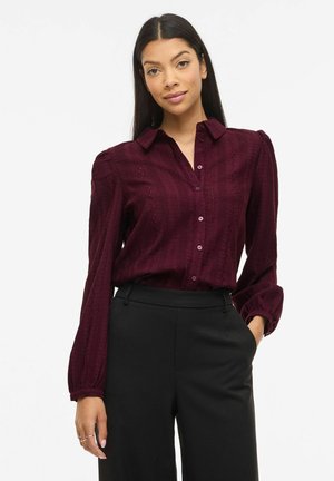 Femme portant un chemisier texturé bordeaux à manches longues avec des boutons et un pantalon taille haute noir, debout avec une main dans la poche.