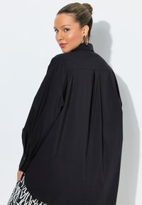 Schwarzes, übergroßes Hemd mit hohem Kragen, langen Ärmeln und Rückenfalten. Glatte Oberfläche, geknöpfte Manschetten und lockere Passform.