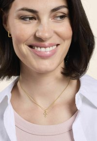 Uśmiechnięta kobieta z krótkimi ciemnymi włosami, nosząca złoty naszyjnik w kształcie krzyża, złote kwadratowe kolczyki, jasnoróżową bluzkę i białą koszulę z otwartym kołnierzykiem.