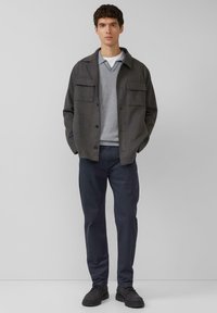 Szara zapinana na guziki kurtka z dwoma kieszeniami na piersi, noszona na paskowanym swetrze, zestawiona z ciemnymi spodniami i czarnymi butami. Prosty, nowoczesny design.