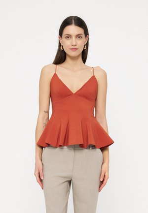 Kobieta ubrana w rdzawe, pomarańczowe, bezrękawnikowe, falbankowe top z cienkimi ramiączkami i beżowe spodnie, stoi na tle białego tła.