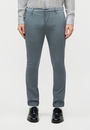 Άνδρας που φοράει ψηλόμεσα στενά παντελόνια μπλε-γκρι, γκρι πουκάμισο με κουμπιά, λευκές κάλτσες και μαύρα δερμάτινα μοκασίνια, στεκόμενος όρθιος.