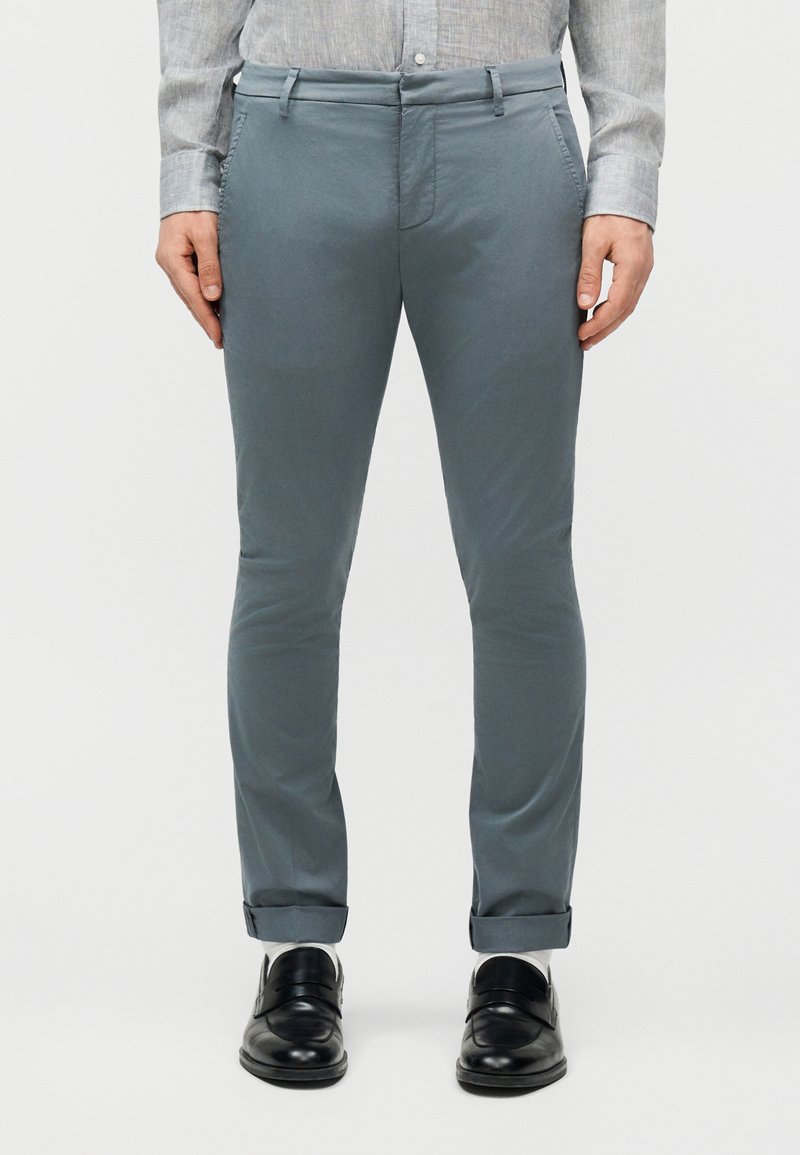 Άνδρας που φοράει ψηλόμεσα στενά παντελόνια μπλε-γκρι, γκρι πουκάμισο με κουμπιά, λευκές κάλτσες και μαύρα δερμάτινα μοκασίνια, στεκόμενος όρθιος.