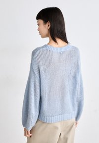 Pull en tricot bleu clair avec une coupe ample, un ourlet côtelé et des épaules tombantes ; il présente une texture transparente et un petit détail de bouton au dos.