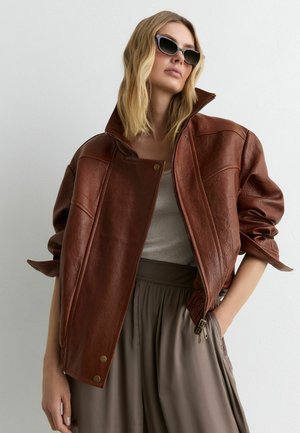 Woman wearing oversized brown leather jacket, gray tank top, loose taupe pants, and black cat-eye sunglasses against white background.