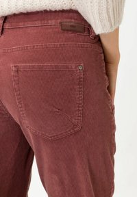 Pantalon en velours côtelé de couleur rose poussiére, avec des nervures verticales, une poche arrière et une petite étiquette logo au-dessus de la ceinture.