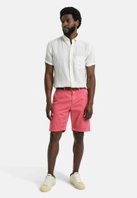 Witte linnen overhemd met korte mouwen en een zak, gecombineerd met roze katoenen shorts en crèmekleurige sneakers met groene accenten. Eenvoudig en casual ontwerp.