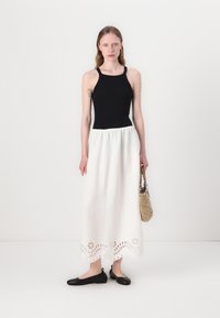 Schwarzes, tailliertes Tanktop, weiße Maxirock mit besticktem floralem Saum, schwarze Ballerinas und Strohtasche. Glatte Textur, legeres Design.