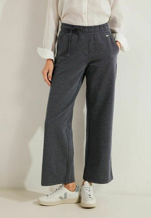 Personne portant un pantalon large gris foncé avec taille à cordon, une chemise blanche boutonnée et des baskets blanches avec des accents bleu clair.