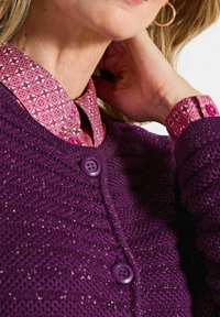 Frau trägt ein pinkes gemustertes Hemd mit einer lila Strickjacke, die glänzende Fäden und passende Knöpfe aufweist, die Hand berührt den Hals, und ein goldener Ohrstecker ist sichtbar.