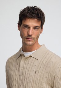 Beżowa koszulka polo w grubym splotem z klasycznym kołnierzem, dwoma guzikami i małym logo na piersi. Nałożona na białą podkoszulkę.