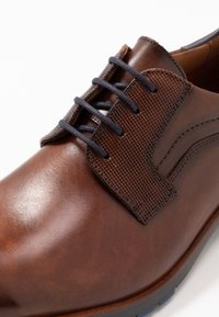 Chaussure en cuir brun avec un panneau en tissu texturé, lacets foncés et une finition lisse avec des détails de couture subtils.