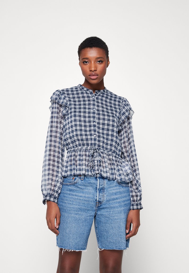 Blusa a quadri blu navy e bianca con maniche a balze e un nodo in vita, abbinata a pantaloncini di jeans sbiaditi con orli sfrangiati.