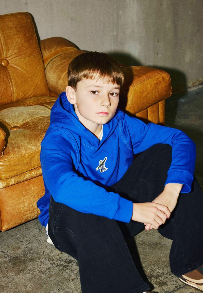 Garçon en sweat à capuche bleu assis par terre, les bras croisés sur les genoux, appuyé contre un fauteuil en cuir marron usé dans une pièce en béton.