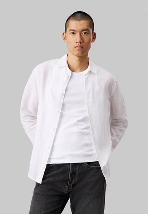 Camisa blanca, ligera, de manga larga con cuello abotonado, superpuesta a una camiseta de tirantes blanca con canalé, combinada con jeans oscuros.