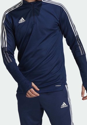 Homme portant un survêtement Adidas bleu marine avec des bandes blanches, des trous pour les pouces sur les manches et un logo sur la poitrine et la cuisse.
