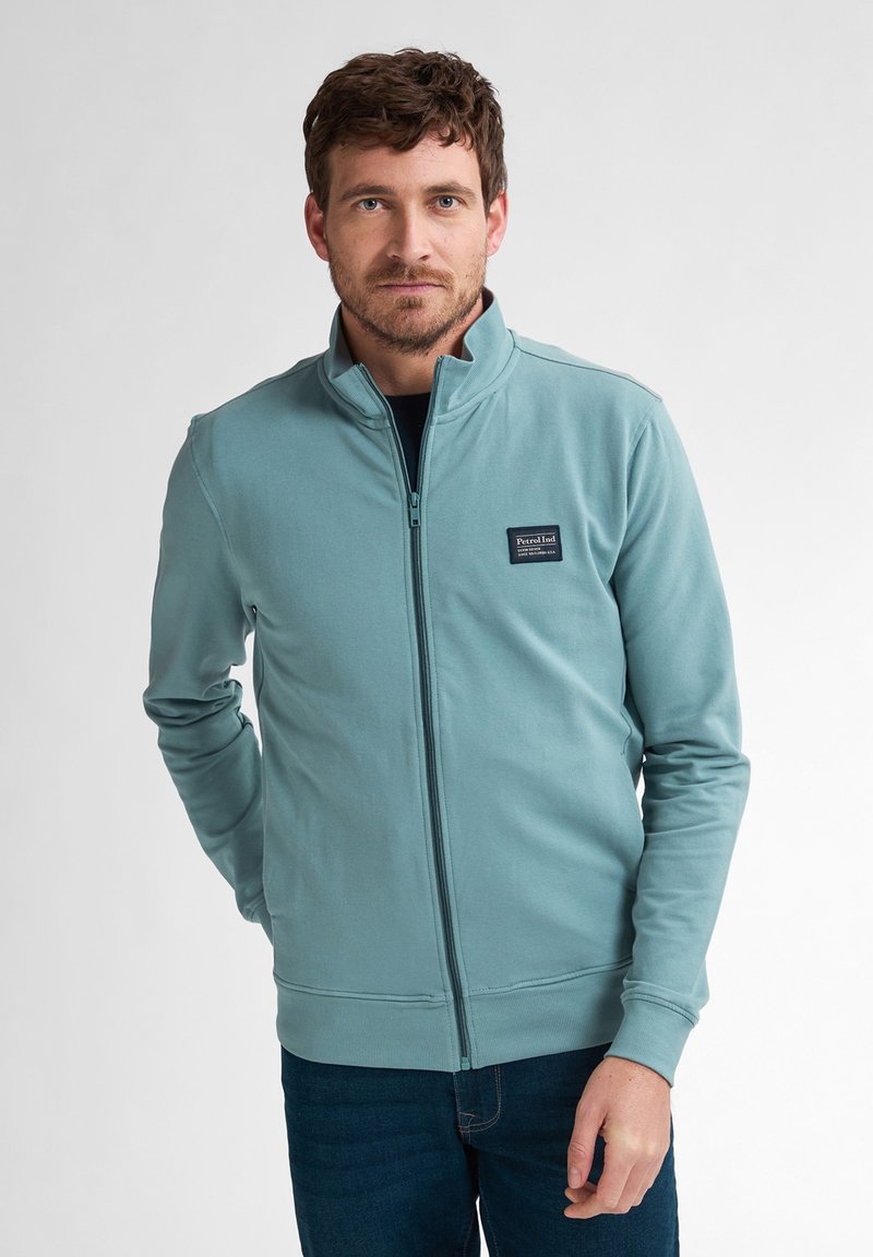 Veste zippée bleu clair avec col haut, fabriquée en matériau doux ; elle présente un patch logo noir et des poignets côtelés. Portée sur un jean foncé.