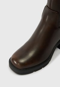 Botim de couro castanho com biqueira quadrada e sola de borracha preta sobre fundo branco, exibindo uma textura suave e detalhes de costura.