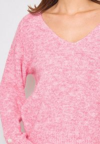 Pull en fluo rose avec un décolleté en V profond, présentant un motif de tricot texturé et des poignets côtelés. Matière douce et chaude pour un maximum de confort.