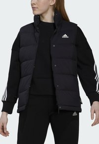 Zwarte puffer vest met een hoge kraag, voorkant met drukknoopsluitingen en Adidas-logo. Gepaard met een zwart longsleeve shirt en strakke broek.