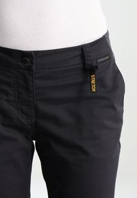 Gros plan d'une personne portant un pantalon noir stretch avec un bouton et une fermeture éclair, et une étiquette visible indiquant "STRETCH" près de la taille.
