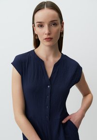 Robe bleu marine à manches courtes avec un tissu texturé, fermeture à boutons, encolure en V et poches latérales, présentant une coupe décontractée et un design subtil.