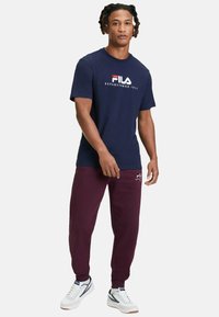 T-shirt en coton navy avec le logo et le texte FILA en blanc et rouge. Pantalons de survêtement bordeaux arborant le logo FILA. Baskets blanches avec des accents bleus.