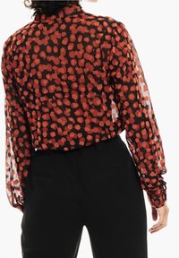 Przezroczysta czarna bluzka z wzorem czerwonych i pomarańczowych pociągnięć pędzla. Długie rękawy z marszczonymi mankietami, w połączeniu z czarnymi spodniami. Widok od tyłu.