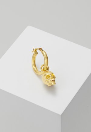 Boucle d'oreille créole en or avec un petit pendentif en forme de crâne détaillé suspendu, présentée sur un bloc géométrique blanc sur un fond gris.