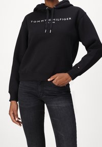 Schwarzer Kapuzensweatshirt aus Baumwollmischung, mit Logo-Druck auf der Brust. Gerippte Bündchen und Saum. Kombiniert mit dunklen Jeans.