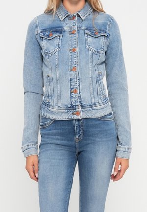 Kvinde iført tætsiddende lyseblå denimjakke med knaplukning og matchende lyseblå denimjeans, stående foran hvid baggrund.