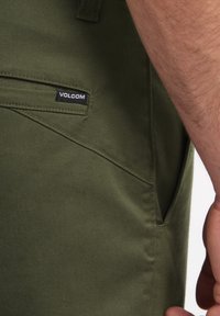 Groene Volcom-broek van gestructureerde stof, voorzien van een zijzak en een merklabel. Het ontwerp is afgewerkt met zichtbare stiknaden.