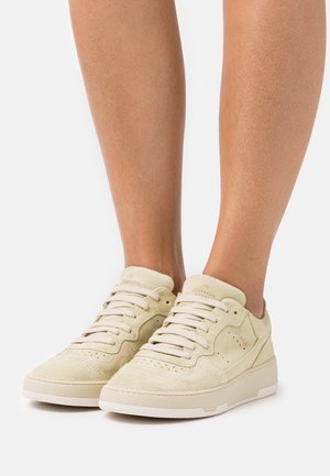 Béžové semišové tenisky s kulatou špičkou, plochým zavazováním na tkaničky, jemnými perforacemi a texturovanou gumovou podrážkou.