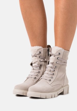 Bottes montantes en daim beige à lacets avec semelles épaisses en caoutchouc et doublure blanche et duveteuse, portées sur des jambes nues sur un fond blanc.