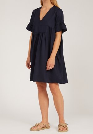 Femme portant une robe bleu marine ample à col en V avec des manches courtes à volants et des sandales en corde beige, debout devant un fond uni.