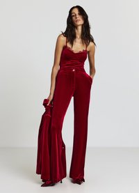 Combinaison en velours rouge avec un corsage ajusté, des détails en dentelle, un pantalon à jambes larges et un bouton à la taille. Portée avec des accessoires assortis.