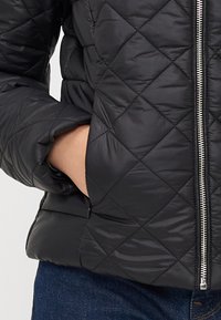 Čierna prešívaná puffer bunda s predným zipsom a bočnými vreckami. Má lesklý povrch a vzor s diamantovým prešívaním.