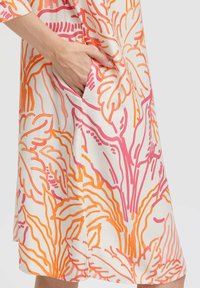 Hand in der Tasche eines weißen Rocks mit auffälligen orangen und pinken Blattmustern, vor einem schlichten weißen Hintergrund.