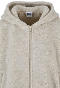 Giacca a cappuccio color crema realizzata in pile morbido con zip a tutta lunghezza e fodera interna. Presenta una superficie morbida e texture.