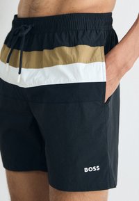 Schwarze Badeshorts mit einem mehrfarbigen horizontalen Streifen (Gold, Weiß) am Bund. Mit Kordelzug und Seitentasche. "BOSS"-Logo am Bein.