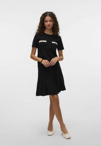 Robe noire à manches courtes, corsage ajusté, jupe évasée, avec des accents horizontaux crème sur la poitrine. Texture tricot lisse.