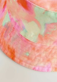 Pūķa cepure ar pasteļtoņu tievā krāsā, kurā ir oranžas, rozā un zaļas tonalitātes. Izgatavota no kokvilnas ar redzamām stitching līnijām gar malām.