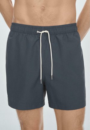 Mann trägt dunkelgraue Schwimmshorts mit weißem Kordelzug, auf Taille und bis zur Mitte des Oberschenkels abgeschnitten, vor einfachem Hintergrund.