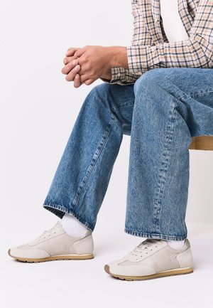 Mann sitzt mit verschränkten Händen, trägt weiße Sneakers, weiße Socken, blaue Jeans und ein kariertes Langarmhemd.