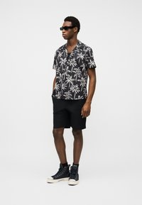 Άνδρας με μαύρα γυαλιά ηλίου, κοντομάνικο πουκάμισο με μοτίβο φοίνικα σε μαύρο, μαύρο σορτς και μαύρα ψηλά αθλητικά παπούτσια, στέκεται μπροστά σε λευκό φόντο.